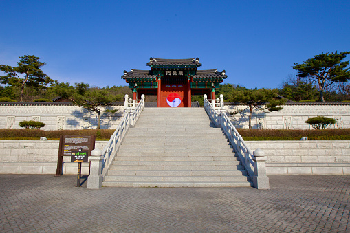 Sangju City, South Korea - March 9th, 2017: The picturesque gate of a Traditional Ritual Hall near the Sangju Museum, featuring concrete stairs, a backdrop of a clear blue sky, a vibrant red door adorned with the Korean Taegeuk symbol, and a traditional hip and gable roof.