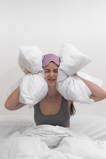 In distress, a woman covers her ears with pillows, unable to sleep due to disruptive neighbors, enduring a throbbing headache and insomnia.