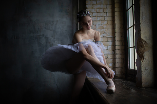 Ballerina adjusts her pointe shoes while standing by the window.