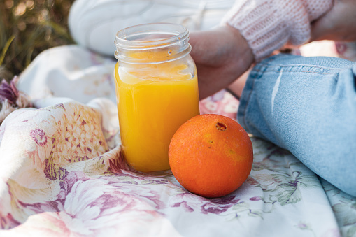 Woman holding with a hand a clean orange juice healthy drink to slim diet having a picnic in the park on a sunny day. Close up of a fresh orange glass bottle. Juice trend and organic food.