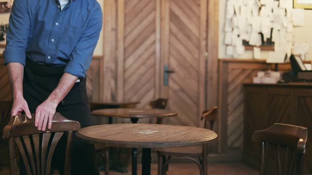 Close up of caucasian food service worker removing chairs from wooden table and placing in order at restaurant. Friendly male in black apron starting workday and waiting for customers indoors.