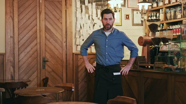 Portrait of handsome bearded waiter adeptly fastening black apron around waist and securing it with bow indoors. Caucasian food service worker preparing for workday in restaurant of retro style.