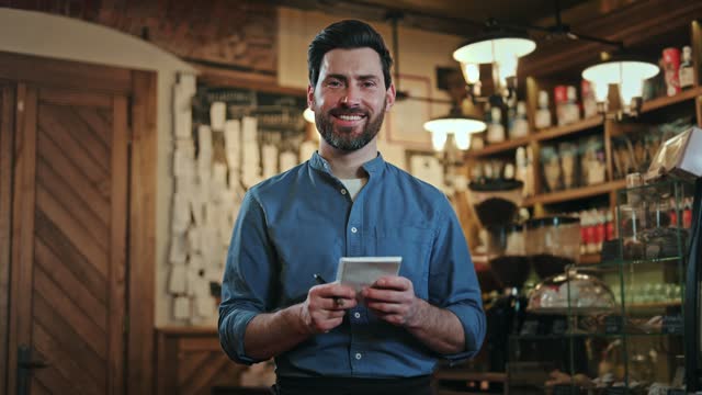 Restaurant staff member with dark hair and beard holding paper notepad while standing at workplace. Caucasian man wearing blue shirt and black apron smiling at camera white taking order notes.