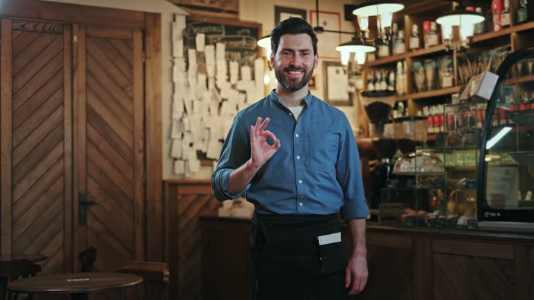 Portrait of handsome service staff showing ok gesture with fingers while expressing positive emotion at cafe space. Caucasian male in shirt and apron making hand sign to agree with something indoors.