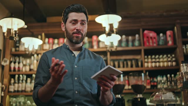Attractive caucasian man dressed in blue uniform smiling and talking pleasantly while taking guest order at stylish cafe. Competent male waiter with beard recording details on paper notepad using pen.