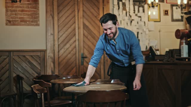 Cheerful employee dressed in apron wiping round tables in eating hall on background of vintage cafe. Handsome bearded man enjoying serving clients and smiling while cleaning furniture during day.