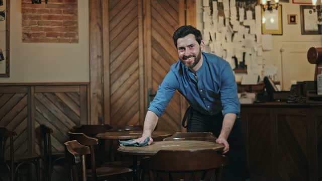 Attractive man cleaning round tables in cafe while smiling and looking at camera. Contented bartender in blue shirt wiping furniture of vintage bistro and preparing to begin working shift in morning.