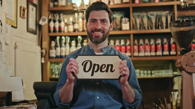 Young entrepreneur opening own restaurant and smiling with happiness while holding wooden sign with writing. Contented worker standing on background of shelves in cafe and welcoming customers.