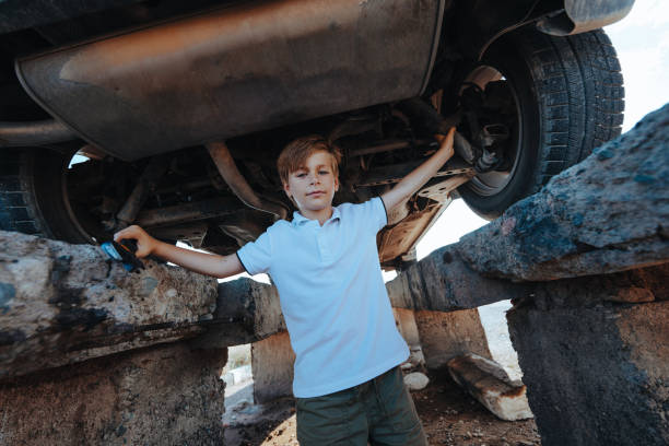 Little boy in blue workwear overall in front of open engine compartment doing some maintenance work at the car