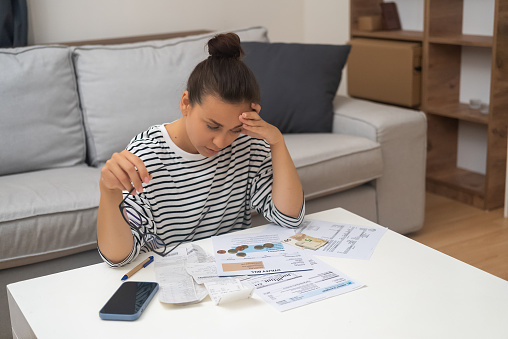 Financial Struggles: Stressed and unhappy, a woman manages household finances, calculates expenses, and seeks relief from debt through online payments.