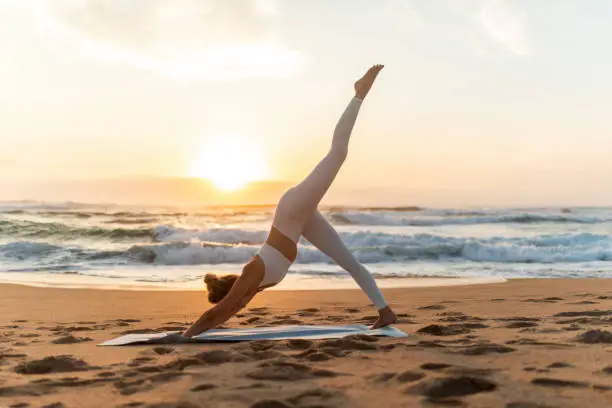 Young woman in sportswear doing yoga exercise with raised leg outdoors, having workout on sea beach at sunset, side view Young woman in sportswear doing yoga exercise with raised leg outdoors, having workout on sea beach at sunset, side view