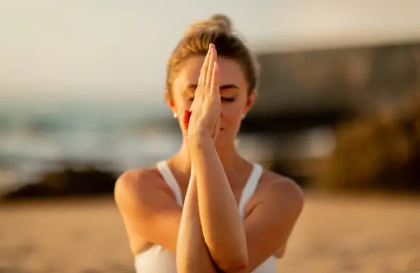 Young european woman deeply engaged in a serene hands-together meditation pose on a sandy beach during a tranquil sunset, closeup shot