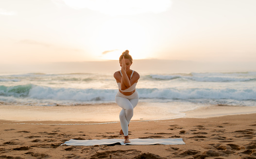 Woman in white activewear focuses deeply as she holds yoga pose, with the sunset casting a warm glow over the crashing waves and sandy shore behind her