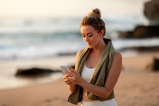 Sporty woman with smartphone resting after outdoors training, lady holding mobile phone and browsing fitness app, standing on sea beach, copy space