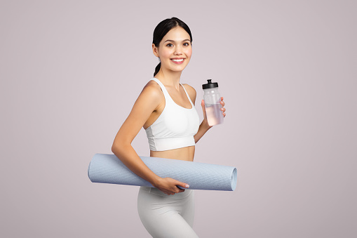 Cheerful caucasian fit woman in sportswear with mat and bottle of water ready for fitness, standing isolated on grey studio background. Sports, body care and aqua balance