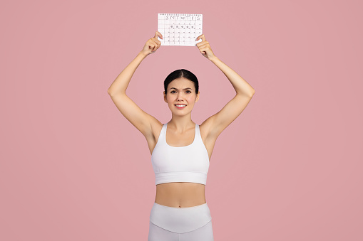 Angry European lady in sportswear, visibly dissatisfied, holds calendar in studio over pink background. The concept touches both athletic routines and menstrual cycle awareness