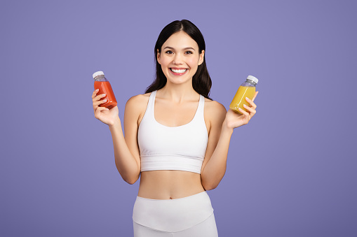 Delighted young woman in white sportswear showcasing two juice bottles, representing healthy choices and lifestyle, against purple background