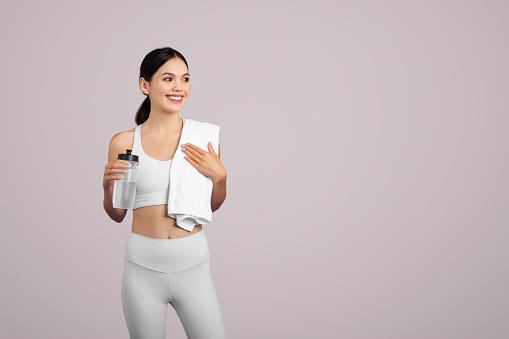 Fit European woman in sportswear holding a water bottle and draping a towel over her shoulder, signaling post-workout on a light background, free space