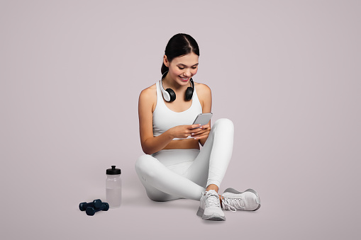 Fit young woman wearing headphones sits relaxed, engaging with her smartphone after workout session, set against light background, full length, free space
