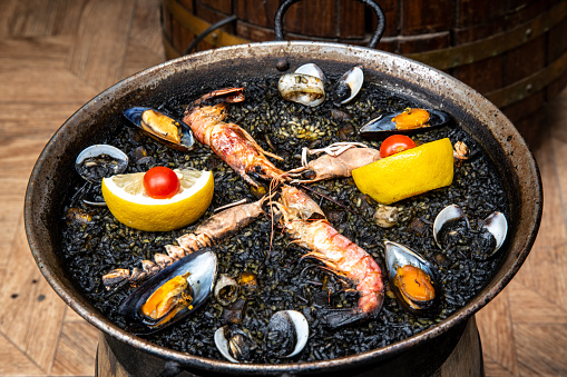 A large paella pan with black rice, lemon, seafood and small tomatoes. You can see scampi, mussels and mussel shells. The pan looks very used.