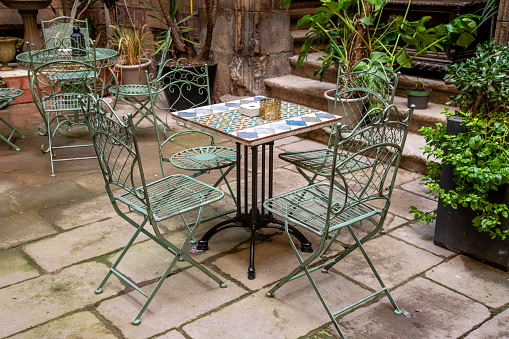 Several metal chairs and tables with artistic elements. The tables are tiled with old tiles and the chairs are wrought iron and green. Everything is set in a cozy courtyard with lots of plants