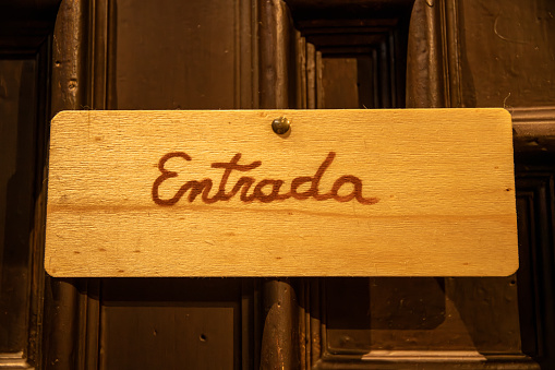 A rectangular wooden sign with the inscription in Catalan: Entrance. The sign is screwed to a brown door.