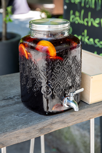 A glass container with a beautiful pattern and a silver lid, as well as a tap with a dark drink in which fruit is floating. The container stands on a rough wooden table.
