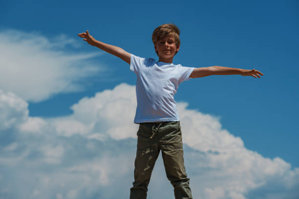 A cheerful boy in a blue shirt sits on a bridge railing, smiling at the camera with lush green mountains and a quiet road behind him.
