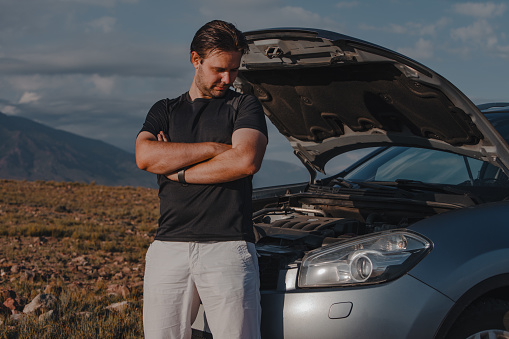 Sad young man standing in front of broken down car on mountains background