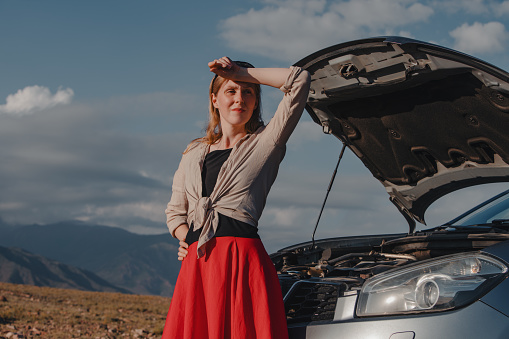Tired young woman standing in front of broken down car on mountains background