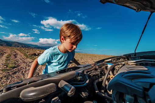 Boy looking into the hood of broken down car on mountains background