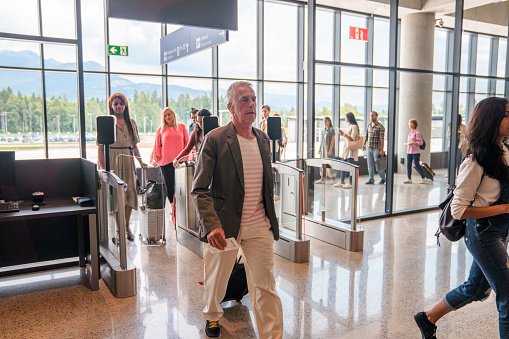 Mature Caucasian male walking past the electronic immigration gates while other people queueing in the background, standing next to a large glass wall. 3/4 length image of the man, wide angle shot.