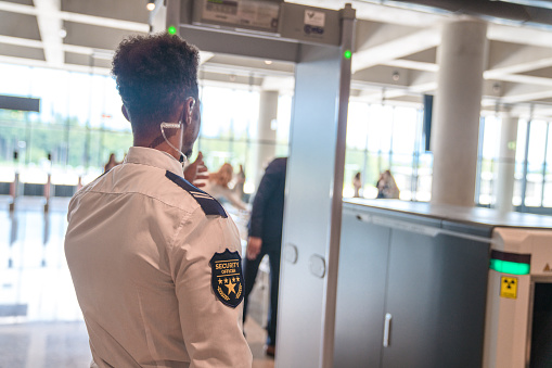 Waist up image of an French-African man working as an airport security officer. Standing next to a x-ray scanner, wearing a uniform and an ear piece and looking in the direction of passengers queueing for the control. Back view.