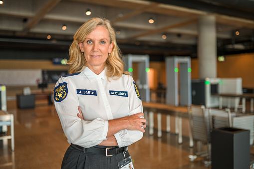 A portrait of an attractive  mid adult Caucasian female security employee posing for a photo in an airport departures lobby, looking at the camera, wearing a uniform shirt with name tag. Waist up shot.
