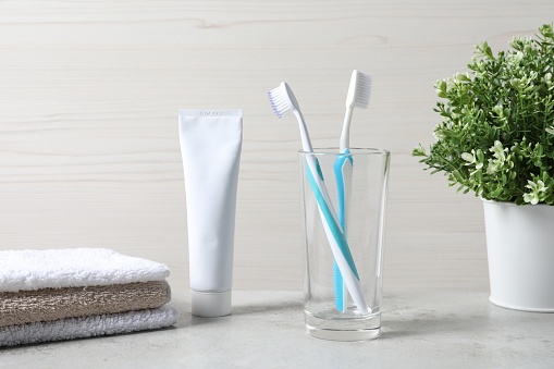 Plastic toothbrushes in glass holder, tube of toothpaste and towels on light grey table