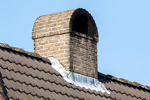 A portrait of a brick chimney on top of a tiled roof during a sunny day. the construction is rounded on top against rain. the inside of the chimney is full of black soot, smoke black or grime.