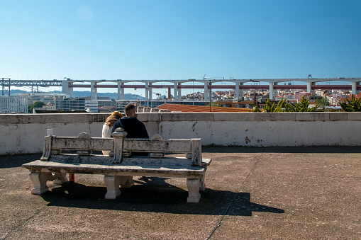 A young couple enjoys a scenic view of Lisbon while seated on a park bench.