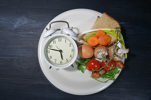 Intermittent fasting, white plate with an alarm clock and partly filled with food on dark blue wood, diet method of eating within a time limit, healthy weight loss, top view from above, copy space