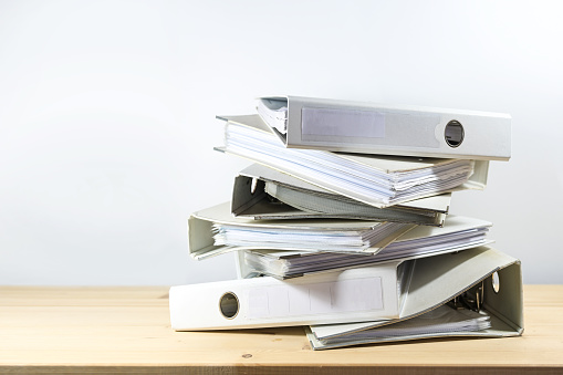 Stack of ring binders on a wooden office desk against a light gray wall, concept of bureaucracy, administration and work overload at the job, copy space, selected focus, narrow depth of field