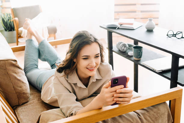 joyful millennial woman relaxing on sofa, engrossed in phone messages, chatting, and enjoying leisure time. content and carefree young female lounging on couch embracing digital communication at home. - news of the world imagens e fotografias de stock