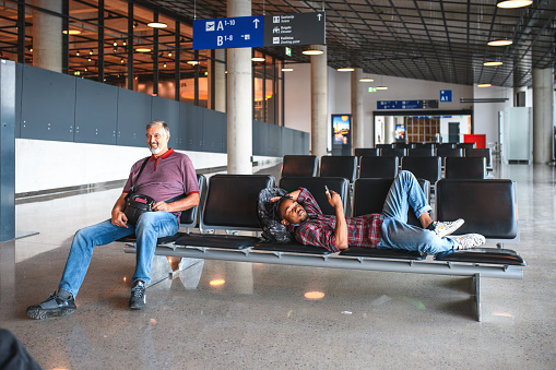 Hispanic male using a smartphone at the airport while lying on the bench in the airport lobby waiting for his flight.