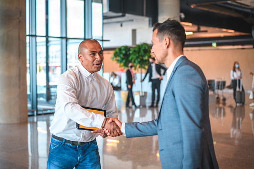 Waist up shot of a Hispanic man shaking hands with a mature Japanese businessman. The Hispanic male is holding a sign with a name written on it.