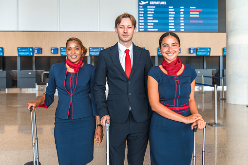 Three quarter length photo of a diverse air crew standing in the airport lobby holding their personal luggage and looking at camera.