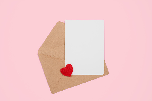 Open craft envelope with empty postcard and heart on pink pastel background, top view with copy space