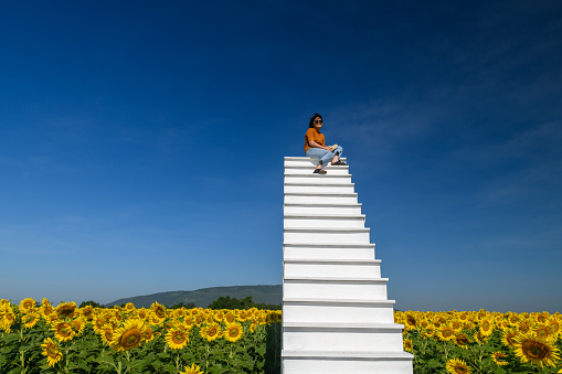 Cute Tourist sitting on the white high stairs with beautiful sunflower field, Lop buri province THAILAND, Travel and relax concept