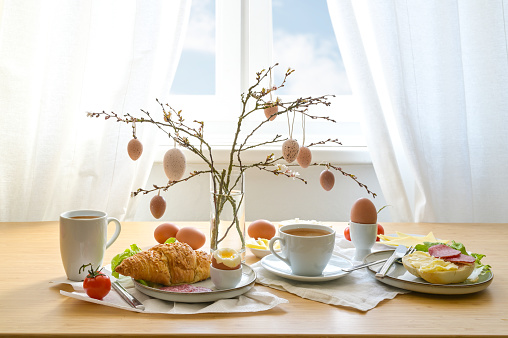 Breakfast with Easter decoration from natural colored eggs hanging on spring branches, coffee, croissant, bread roll and more on a wooden table, copy space, selected focus, narrow depth of field
