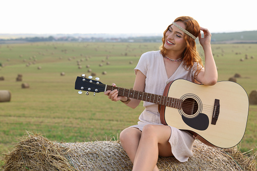Beautiful hippie woman with guitar on hay bale in field, space for text