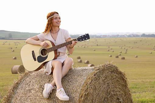 Beautiful hippie woman playing guitar on hay bale in field, space for text