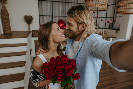 Young man making selfie while embracing his beautiful girlfriend holding a flower bouquet at decorated home