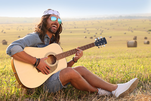 Handsome happy hippie man playing guitar in field